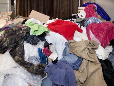 Large messy pile of household items, clothes, boxes, hangers, bubble wrap, toilet paper, pants, shirts, dresses with curtains and mirror in background