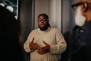 A man has a serious expression as he talks to onlookers. He uses his hands to gesture as he explains and discusses a matter with his coworkers / audience.