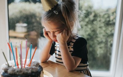 little girl wearing birthday hat look grumpily at her birthday cake