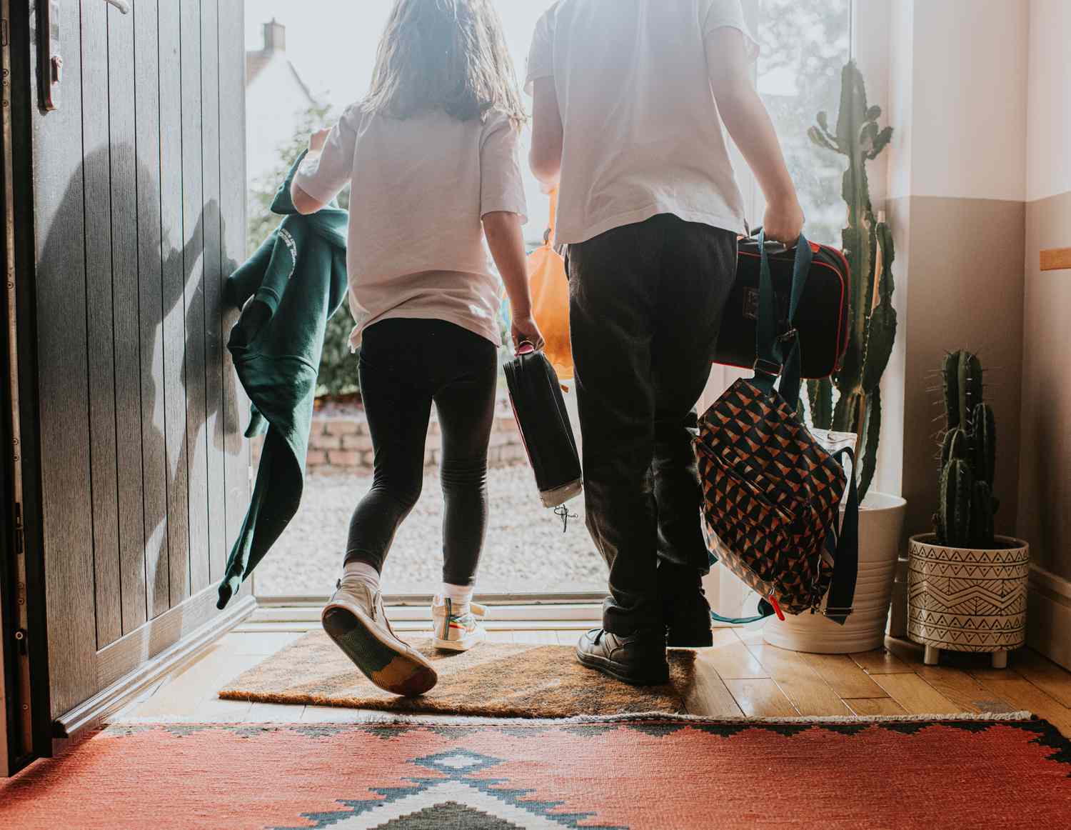 Two young children exit a doorway at the same time, holding backpacks and lunch bags and wearing a school uniform. Image depicts a morning and the school day ahead.