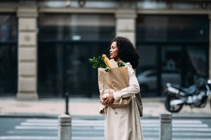 Woman with curly hair carrying a paper grocery bag crossing a street in an urban setting. She portrays a casual, everyday moment.