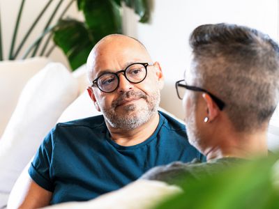 A man listens to his partner speak as they sit together face to face on their couch at home.