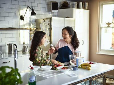 Woman holding yoghurt on spoon towards girl, laughing, threatening to smear food on daughter's face, messing about, having fun, playing, bonding