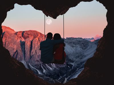 Couple on swing contemplating the mountains in a romantic view with heart shape. Composition of the Alps mountains during sunset with full moon and couple on swing from a heart shape cave.