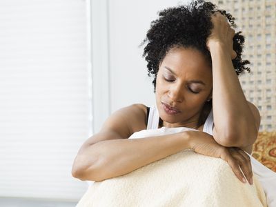 A woman with black curly hair sits with her knees to her chest. She holds her head in her arm and closes her eyes in distress.