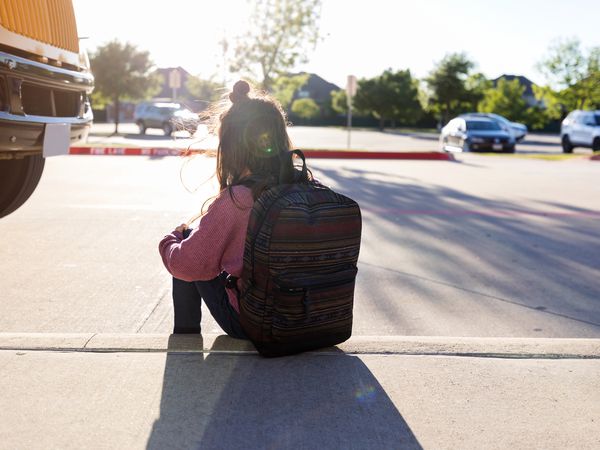 schoolgirl with a backpack hugs her knees and sits on the curb by herself at a bus stop