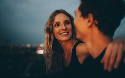 Smiling woman with arm around looking at man while sitting on terrace at dusk 