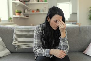 Frustrated young woman suffering from the headache while sitting on the sofa at home with an expression of being unwell, with eyes closed.