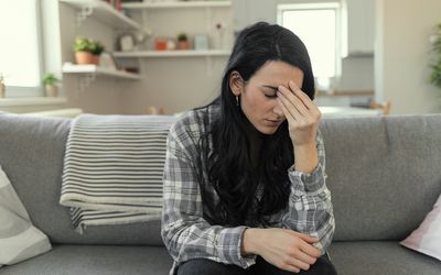 Frustrated young woman suffering from the headache while sitting on the sofa at home with an expression of being unwell, with eyes closed.