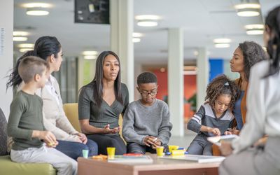 Women and kids sit around a table in a colourful facility and discuss.