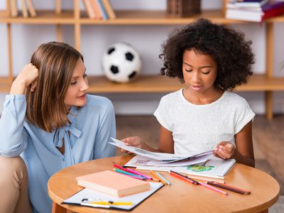 Positive psychologist looking at african american girl with pictures at coffee table on blurred background