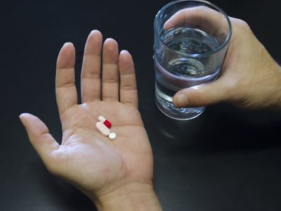 Hands holding three pills and a glass of water against black background, copy space