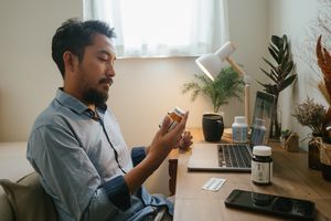 Asian man looking at pill bottle and listening to women doctor in consultation medicine to video conference at home.