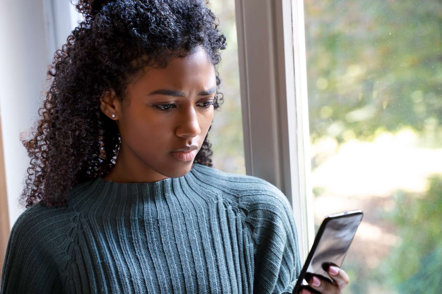 African American female stares outside her window looking cellphone.