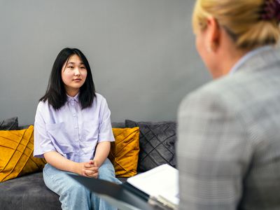 A serious teenage girl gestures as she sits on a couch in her school counselor's office and talks to her unrecognizable counselor. The counselor takes notes on a clipboard.