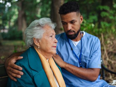 Caregiver man helping and supporting senior woman sitting outdoors in park.