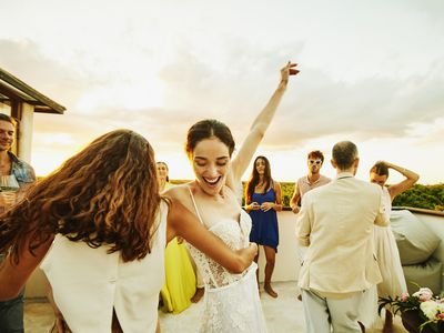 Medium wide shot of smiling bride and friend dancing during party on rooftop deck after wedding at tropical resort