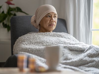 An older woman wearing a head scarf sits at home and looks contemplatively out the window.