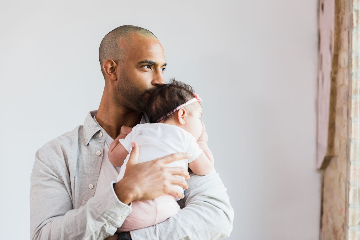 father looks out the window in his home as he carries his baby daughter.