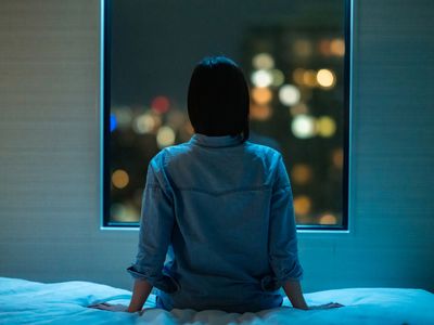 Rear view of woman sitting alone on bed in room and looking through window at night