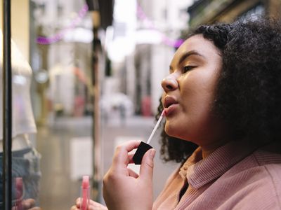 Young woman with curly hair applying lip gloss in front of store window