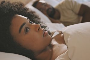 Worried female laying in bed with her husband looking anxious and concerned while thinking of her relationship issues. A man sleeping while his wife lays awake at night feeling depressed and troubled