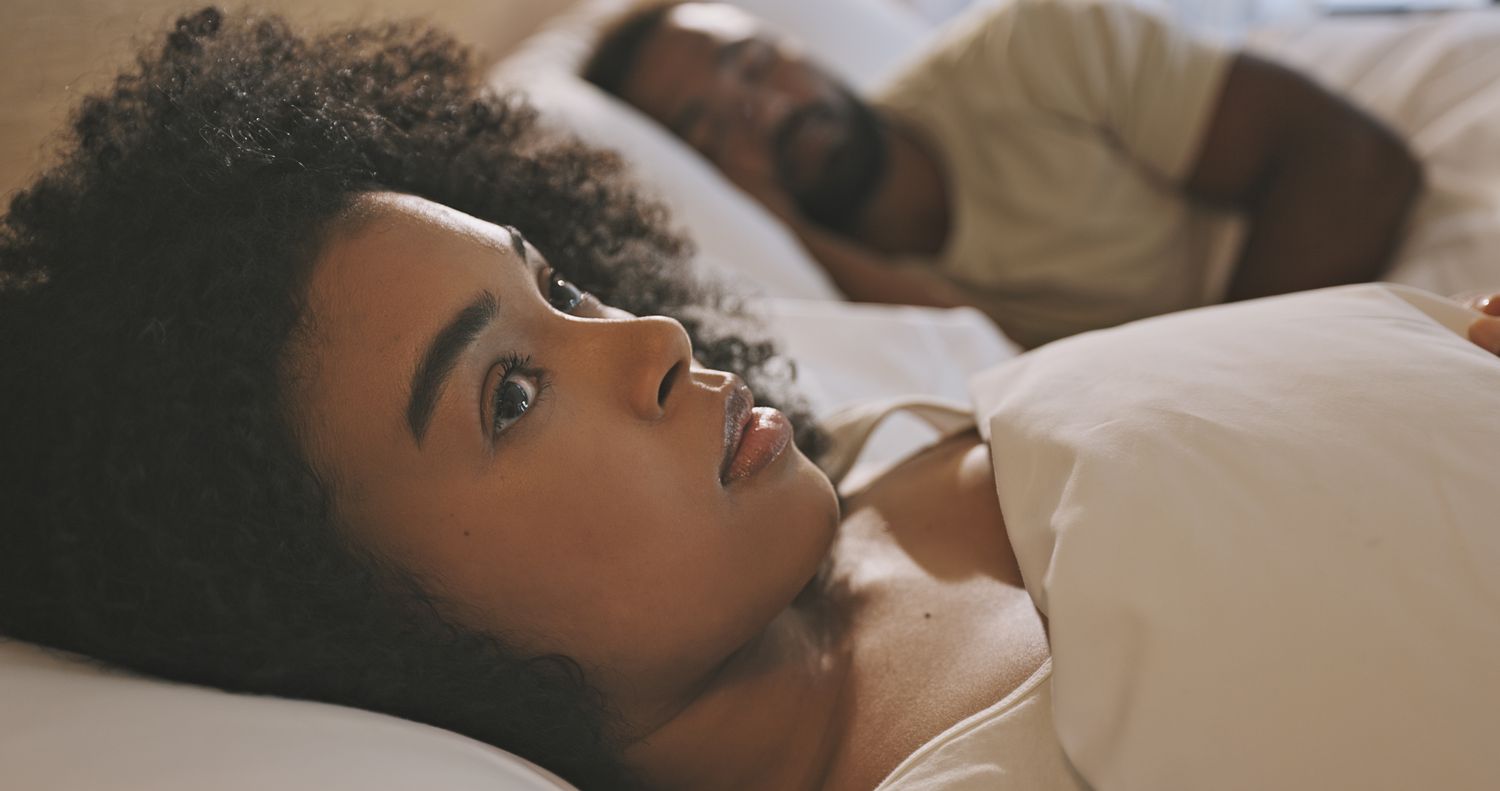 Worried female laying in bed with her husband looking anxious and concerned while thinking of her relationship issues. A man sleeping while his wife lays awake at night feeling depressed and troubled