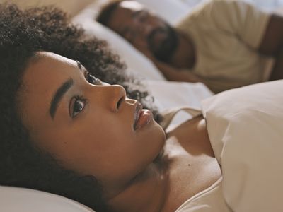 Worried female laying in bed with her husband looking anxious and concerned while thinking of her relationship issues. A man sleeping while his wife lays awake at night feeling depressed and troubled