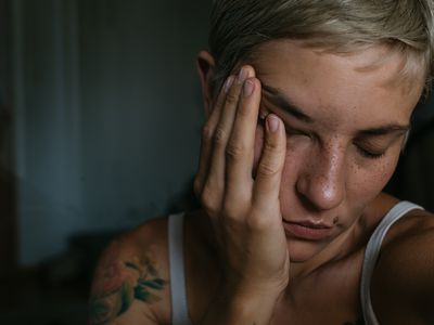 A horizontal closeup portrait of a young woman in natural lighting on a dark background holding hand to face with short dyed blond hair with closed eyes and skin texture and freckles makeup with a birthmark on face and flower tattoos looking tired or having a headache