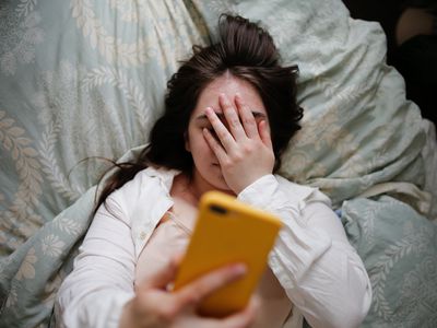 Plus size young woman in home bedroom on bed with phone. Woman covering her eyes with her hand