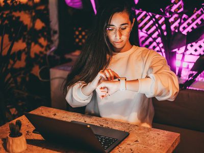 Young millennial woman in smart glasses is working in modern neon cafe with laptop at night, using wireless devices. The female is checking her health data with smart watch or replying a message instantly. Female investor and trader monitoring stock market