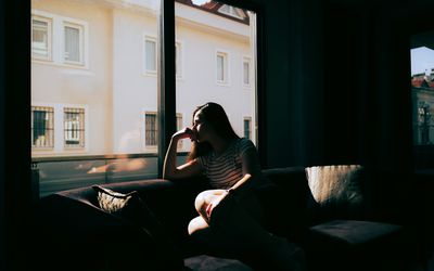 A tired and depressed Mediterranean woman is sitting on the sofa near the window at home, feeling exhausted and devastated. Emotional stress, burnout, and relationship difficulties that need to be taken care of