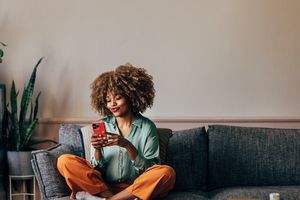 A smiling elegant African-American female using her smartphone while sitting on the cozy sofa in the living room.