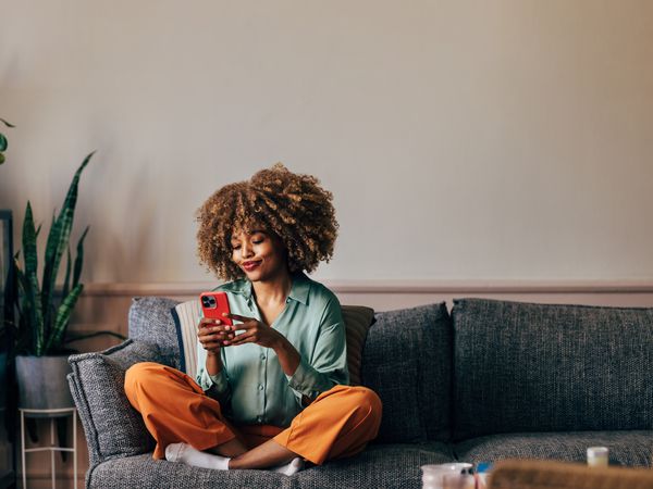 A smiling elegant African-American female using her smartphone while sitting on the cozy sofa in the living room.