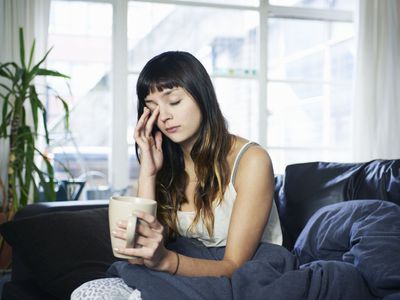 woman with closed eyes touching her face, sitting on sofa with coffee cup in hand