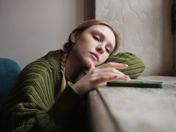 Upset sad millennial girl sitting by window looking at mobile phone waiting call from boyfriend