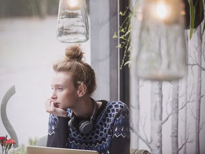 young woman with headphones around her neck and chin resting on hand, looking out cafe window