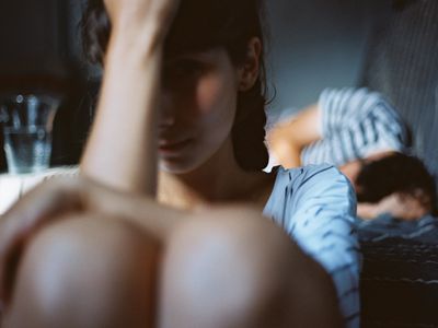 Man sleeping in bed with an unhappy woman sitting on the floor next to him