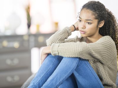 A teenage girl of African descent is indoors in a bedroom. She is wearing casual clothing. She is sitting on her bed and looking sad.