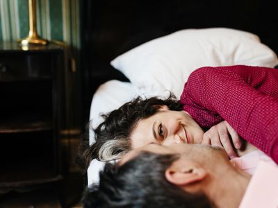 A mature, loving couple is looking at each other lying on a hotel bed. They seem to be in love as the woman smiles gladly at her partner. In the background, dark furniture and a green wallpaper can be seen.