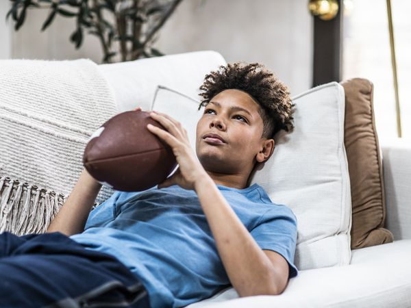 Black teenager lying on bed holding football while gazing into the distance