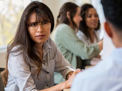 A serious young female creative professional sits at a conference table during a staff meeting next to an unrecognizable coworker. She frowns in disbelief as she listens to him.