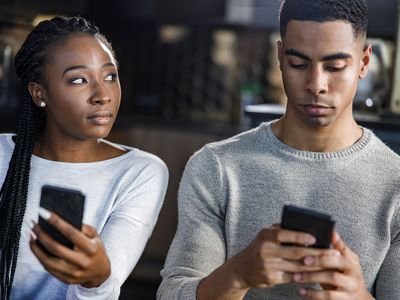 Suspicious Black woman peeking into her boyfriend's cell phone while he is text messaging to someone.