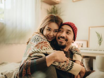 Happy young family couple relaxing talking laughing holding cups drinking coffee tea sitting on bed together in bedroom, loving husband and wife bonding enjoying pleasant conversation at home