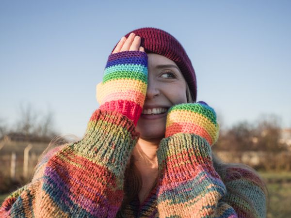 Smiling woman wearing rainbow gloves covering face with hands on sunny day